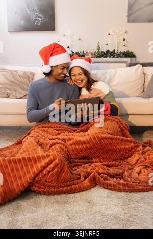 Couple diversifié portant des chapeaux de Père Noël, se penchant près sur un tapis, partageant tablette et couverture rouille-orange. Cosy, intime, festif, vivant Banque D'Images