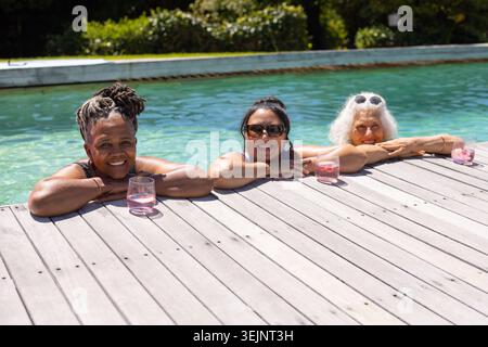 Diverses amies féminines réchauffant la peau au soleil, appuyées sur la terrasse en bois à la piscine, sirotant des boissons roses. Piscine, trio, lunettes de soleil, turquo Banque D'Images