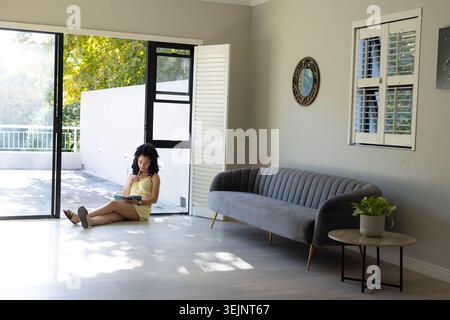 Femme assise sur le sol près de la porte en verre, tenant un cahier et un stylo, portant une robe jaune. Ensoleillé, patio, minimaliste, serein, suite Banque D'Images