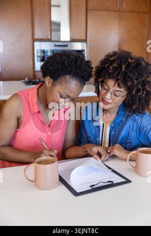 Diverses amies féminines assises au comptoir de cuisine examinant et signant le formulaire sur le presse-papiers avec un stylo. Collaboration, paperasse, mu Banque D'Images