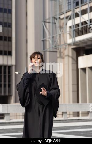 Femme dans la vingtaine en robe académique debout sur la plaza tenant le chapeau, parlant sur smartphone. Robe, mortarboard, diplômé, Terra Banque D'Images