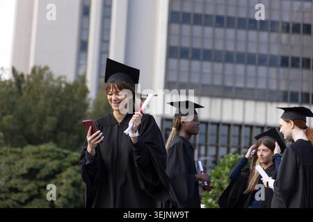 Diplômées d'université en robes noires détenant des diplômes avec des rubans rouges et téléphone sur la pelouse du campus. Étudiants, graduation, mortier Banque D'Images
