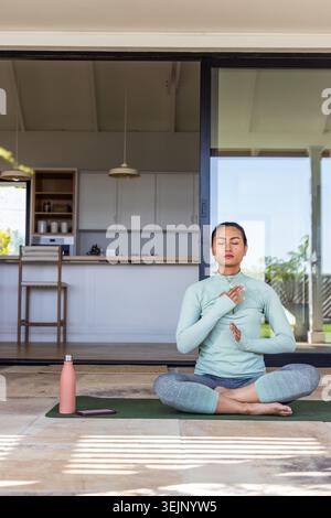 Femme asiatique dans le top vert menthe assis méditant sur le tapis vert à la porte patio avec bouteille rose. Pleine conscience, sérénité, ensoleillé, dehors Banque D'Images