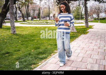 Jeune femme noire marchant dans le parc envoyant un message texte Banque D'Images