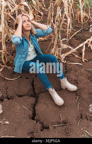 Triste femme fermière tenant sa tête dans ses mains dans un champ de sol fissuré sec à côté de tiges de maïs fanées Banque D'Images