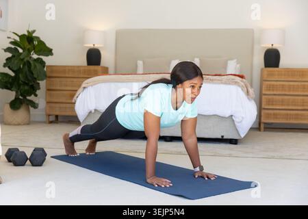 Femme afro-américaine exécutant planche sur tapis de yoga dans la chambre en vêtements de sport avec haltères Banque D'Images