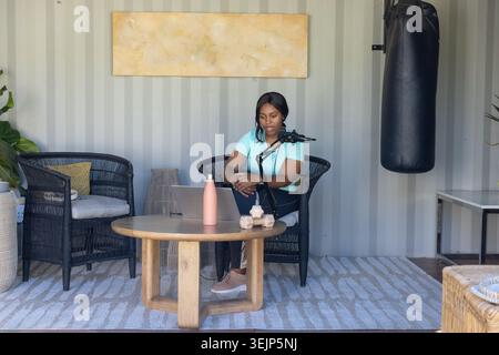 Femme africaine adulte podcasting sur micro d'ordinateur portable dans le studio assis sur une chaise en osier près du sac de punching Banque D'Images
