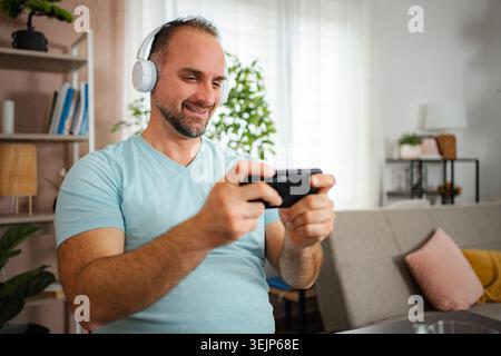 Homme portant des écouteurs jouant à un jeu vidéo sur smartphone à la maison Banque D'Images