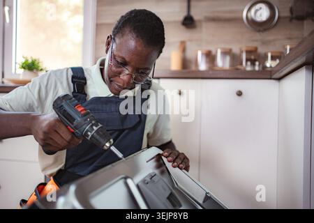 Technicien réparant lave-vaisselle à l'aide d'un tournevis électrique dans la cuisine Banque D'Images