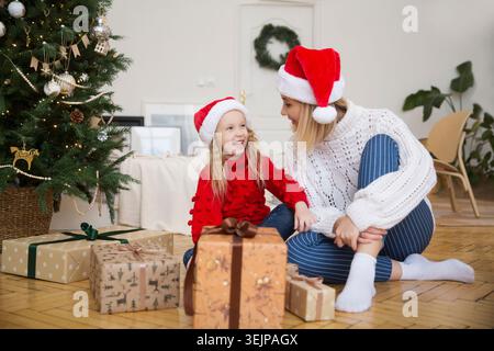 Maman et fille enfant dans un chapeau Santas avec des cadeaux de Noël à la maison près de l'arbre de Noël. La famille rencontre Noël ensemble Banque D'Images