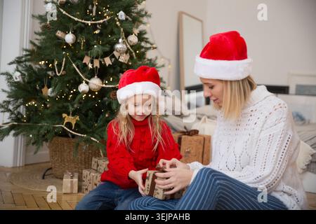 Maman et fille enfant dans un chapeau Santas avec des cadeaux de Noël à la maison près de l'arbre de Noël. La famille rencontre Noël ensemble Banque D'Images