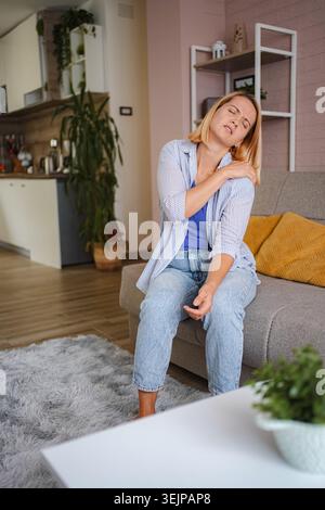 Femme souffrant de douleurs à l'épaule en étant assise sur un canapé à la maison Banque D'Images