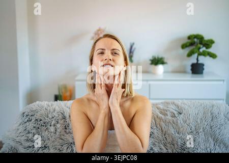 Femme adulte moyenne appliquant un hydratant sur le cou assis sur le canapé à la maison Banque D'Images