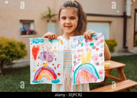 Fille souriante montrant ses dessins colorés dans le jardin Banque D'Images