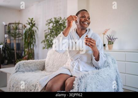 Femme noire souriante appliquant le sérum sur le visage à la maison Banque D'Images