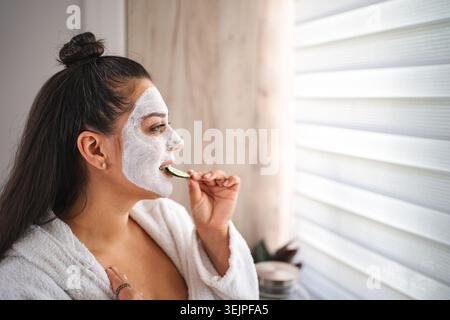 Jeune femme portant un peignoir mangeant du concombre avec un masque facial regardant par la fenêtre Banque D'Images
