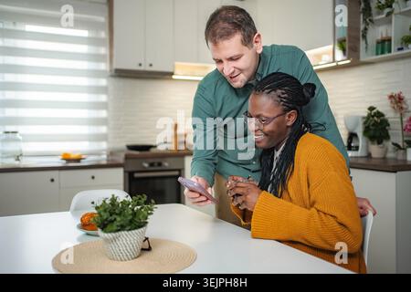 Couple multiracial partageant le contenu de smartphone dans la cuisine Banque D'Images