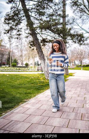 Jeune femme noire marchant dans le parc vérifiant smartphone Banque D'Images