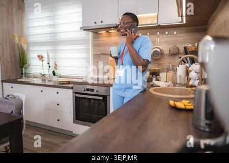 Infirmière souriante parlant au téléphone et buvant du café pendant sa pause dans la cuisine Banque D'Images