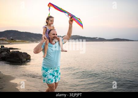 Père portant sa fille sur les épaules tenant le cerf-volant au coucher du soleil Banque D'Images