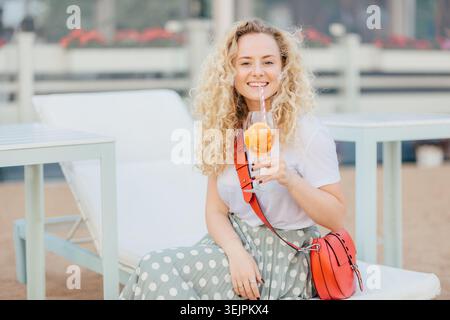 Femme blonde heureuse avec les cheveux clairs bouclés, tient le cocktail orange frais, étant dans l'esprit élevé, habillé de façon décontractée, pose en plein air sur Banque D'Images