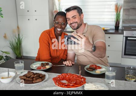 Couple interracial prenant un selfie tout en déjeunant dans la cuisine Banque D'Images
