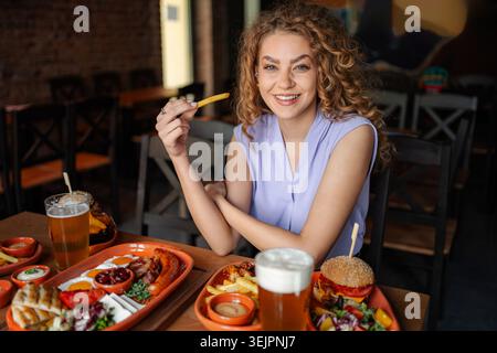 Jeune femme mangeant des frites dans un restaurant avec une table pleine de nourriture et de bière Banque D'Images