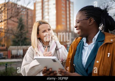 Diverses femmes amies parlant rire en utilisant la tablette à l'extérieur Banque D'Images