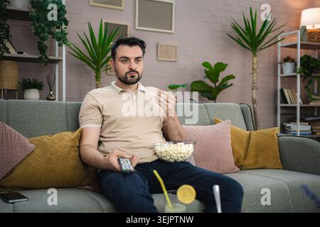 Jeune homme regardant la télévision tout en mangeant du pop-corn sur le canapé Banque D'Images