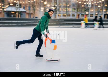 Portrait de l'homme barbu heureux porte des vêtements à la mode utilise l'aide de skate pour garder l'équilibre sur la glace, apprécie les week-ends dans le cercle de Banque D'Images