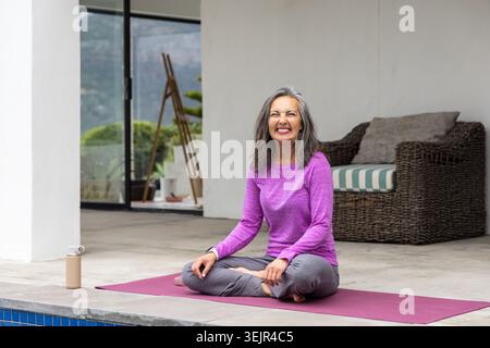 Femme senior assise sur les jambes croisées sur le tapis de yoga sur le patio, portant le dessus violet avec bouteille d'eau à proximité. Plein air, sérénité, bien-être Banque D'Images