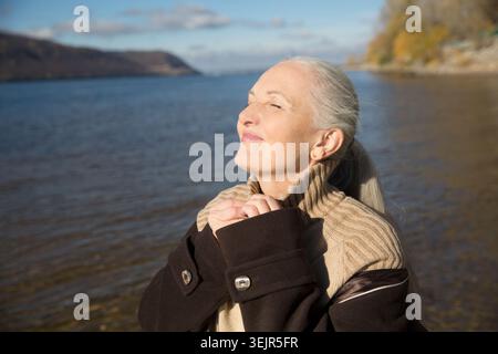 Femme âgée avec les cheveux gris portant un manteau brun sur la rive de la rivière. Vie lente. Profiter des petites choses. promenades en aute Banque D'Images
