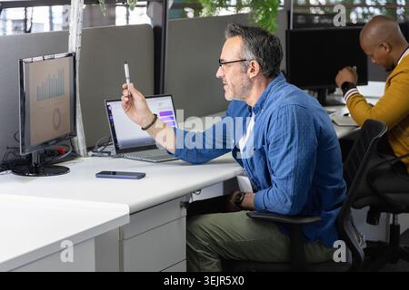 Divers collègues masculins travaillant dans le bureau, homme tenant le stylo vers le moniteur avec le téléphone portable. Espace de travail, travail d'équipe, ergonomie, journée Banque D'Images