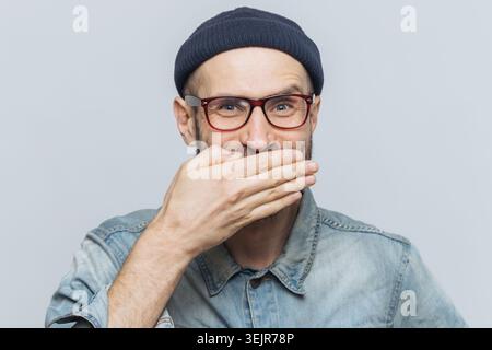 Homme barbu surpris dans des lunettes couvrant la bouche avec la main isolée sur fond gris Banque D'Images