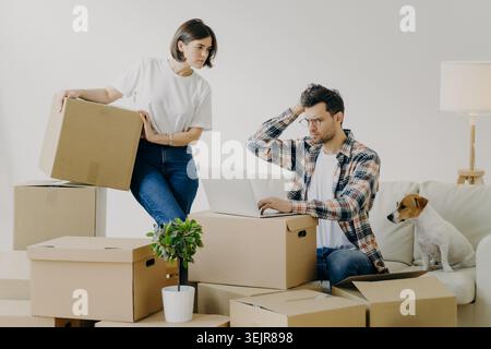 Jeune couple déménageant la maison avec des boîtes et ordinateur portable tandis que le chien est assis sur le canapé Banque D'Images
