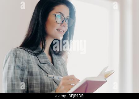 Femme d'affaires écrivant dans un cahier par fenêtre tout en planifiant le travail dans le bureau Banque D'Images