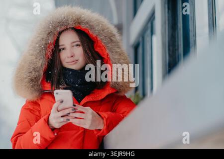 Jeune femme utilisant un smartphone à l'extérieur en hiver portant une parka rouge et une capuche en fourrure Banque D'Images
