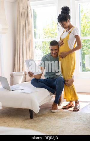 Couple afro-américain lisant le tableau à la maison, femme en salopette de moutarde tenant la bosse, espace de copie. Enceinte, partenaires, livingroo Banque D'Images