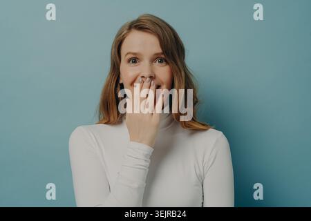 Surprise jeune femme couvrant la bouche avec la main sur fond bleu montrant une réaction choquée Banque D'Images