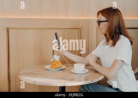 Jeune femme utilisant smartphone dans le café avec des écouteurs ayant le café et croissant petit déjeuner Banque D'Images