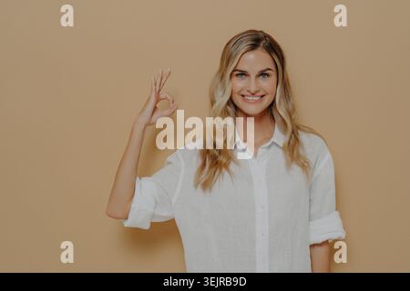 Sourire jeune femme montrant OK geste de main sur fond beige portant chemise blanche Banque D'Images