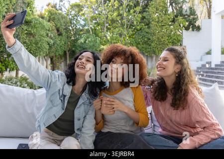 Diverses amies féminines posant pour selfie sur un canapé extérieur blanc, tenant un smartphone et une tasse rose. Trio, copines, patio, canapé Banque D'Images