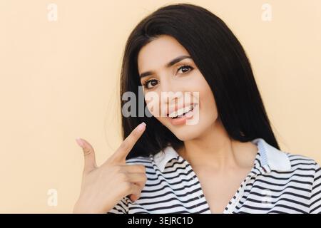 Jeune femme souriant avec des appareils dentaires pointant vers les dents sur fond Beige Studio Banque D'Images