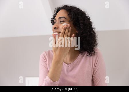 Souriant adulte non binaire appliquant de la crème blanche à la joue gauche portant des anneaux de chemise rose chaîne dans le grenier. Soin de la peau, hydratant, self Banque D'Images