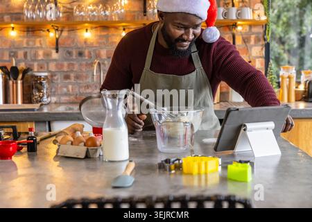 Homme afro-américain portant chapeau de Père Noël et tablier mélangeant la pâte et consultant tablette dans la cuisine. Pâtisserie, maison, confortable, biscuits, re Banque D'Images