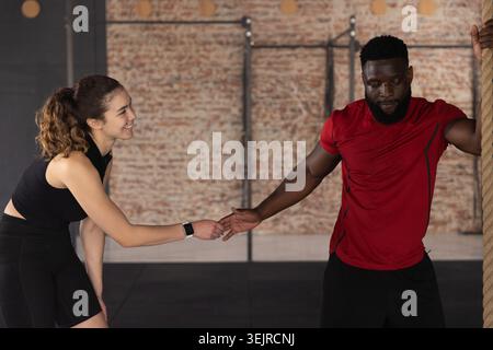 Couple diversifié portant des vêtements athlétiques dans la salle de gym, homme tenant la corde, femme atteignant smartwatch souriant. Duo, fitness, entraînement, brique Banque D'Images