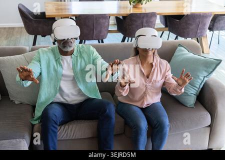 Couple senior assis sur un canapé gris portant des casques blancs VR et atteignant le salon virtuel. Canapé, maison, lunettes, immers Banque D'Images