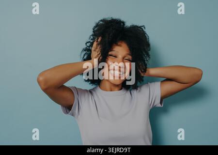 Heureuse jeune femme noire riant avec les mains dans les cheveux sur fond bleu dans le Portrait studio ludique Banque D'Images