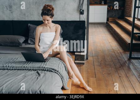 Détendu jeune femme utilisant un ordinateur portable sur le lit à la maison enveloppé dans une serviette dans une chambre loft moderne Banque D'Images