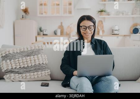 Femme souriante travaillant sur ordinateur portable à la maison assis sur canapé dans le salon confortable pendant le travail à distance Banque D'Images
